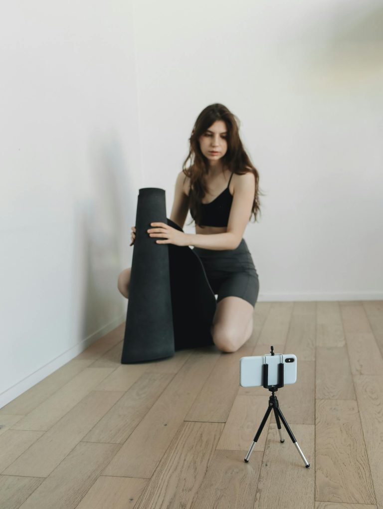 A young woman prepares her yoga mat in a minimalist room for an online workout session using her smartphone.