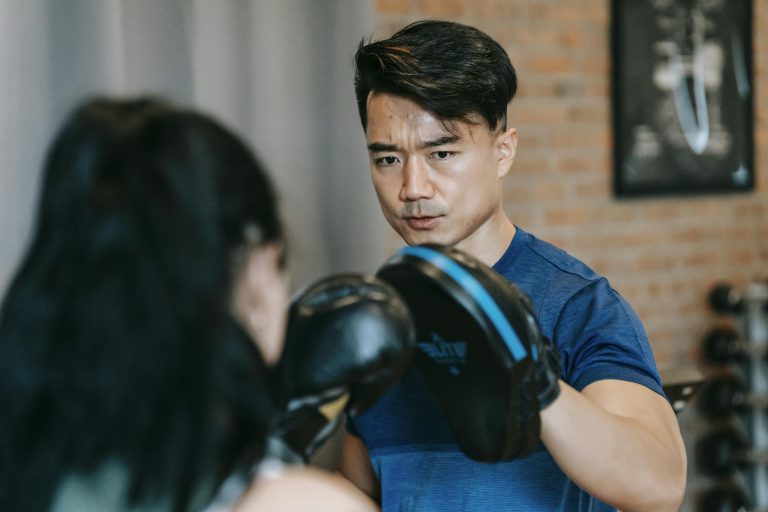 Woman in boxing gloves punching paw of concentrated Asian personal instructor during workout in gym